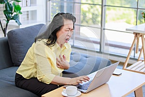 Asian old senior pensioner chubby fat housewife sitting on cozy sofa open mouth wide shocking holding hand on chest having heart
