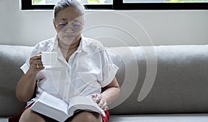 Asian older woman holding cup of coffee and reading a book beside the window after get up in morning