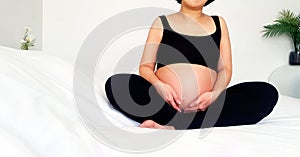 Asian pregnant woman in black undershirt sitting on white bed and touching her belly in the bedroom with copy space