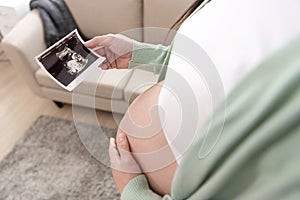 Asian pregnant woman hand holding ultrasound image on belly at home. Pregnancy, maternity, preparation and expectation concept