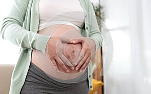 Asian pregnant woman hands making heart gesture on belly at home. Pregnancy, maternity, preparation and expectation concept