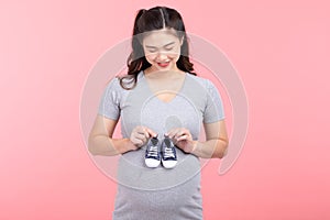 Asian pregnant woman holding baby sneakers for baby newborn isolated on pink background. Pregnant woman packing baby stuff ready