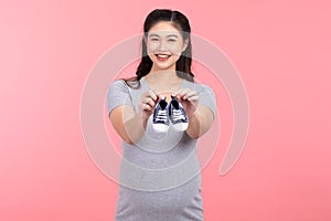 Asian pregnant woman holding baby sneakers for baby newborn  on pink background. Pregnant woman packing baby stuff ready