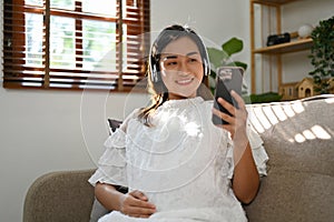 Asian pregnant woman listening to music through headphones, using her mobile phone