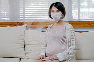 Asian pregnant woman with mask sit on sofa in living room and look to camera with concept protection from coronavirus or toxicant