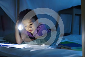 Asian preschool boy child to make a camp to play imaginatively and reading book by flashlight  in living room at home