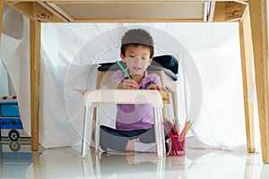 Asian preschool boy drawing in paper while lying in a blanket fort in living room at home for perfect hideout away from their