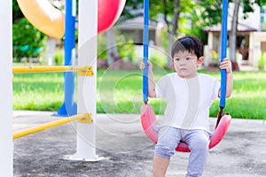 Asian preschool little child boy sitting on red swings  lonely or sad gestures unhappy. A scared little child is 2 years old.