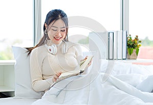 Asian pretty female teenage girl in turtleneck sweater wearing headphones smiling reading a book and sitting on white clean sheet