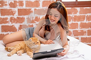 Asian pretty girl lie on bed and fun together with orange cat that look enjoy to tablet
