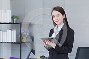 Asian professional business young woman in black suit smiles happily stand and look at the camera while she works and holds tablet