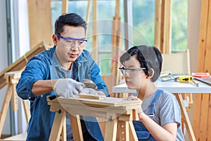 Asian professional carpenter engineer dad teaching young boy son in jeans outfit with gloves safety goggles nailing steel hammer