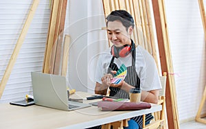 Asian professional cheerful happy male carpenter worker staff in apron with earphones sitting smiling holding video call via