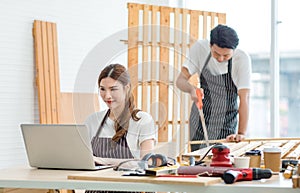 Asian professional female carpenter worker in apron sit typing data information on laptop computer while male colleague standing