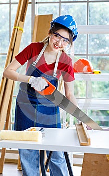 Asian professional female engineer architect foreman labor worker wears safety goggles hard helmet and jeans apron using handsaw