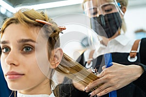 Asian professional female stylist cutting Caucasian woman`s hair in salon. Hairdresser wear mask and face shield to prevent from