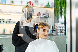 Asian professional female stylist cutting Caucasian woman`s hair in salon. The man wearing mask and face shield to prevent from co