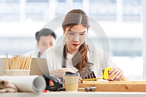 Asian professional focused female carpenter worker staff in apron sitting holding using measuring tape measure wooden sticks on