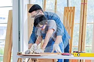 Asian professional male carpenter woodworker engineer dad in jeans outfit with safety gloves goggles helping teaching young boy