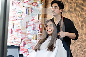 Asian professional male hairstylist combing and using scissors cutting woman`s hair in salon. Hairdresser talking to young girl photo
