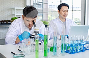 Asian professional male scientist researcher in white lab coat and rubber gloves sitting using microscope inspecting quality of
