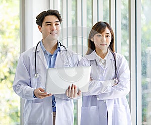 Asian professional young male and female intern doctor in white lab coat with stethoscope standing crossed arms holding laptop