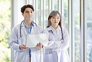 Asian professional young male and female intern doctor in white lab coat with stethoscope standing crossed arms holding laptop
