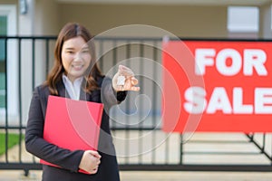 Asian real estate agent or realtor woman smiling and holding red file with showing house key in front of house