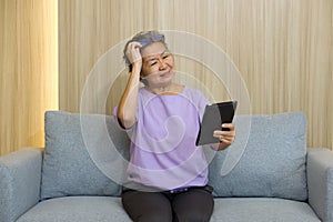 Asian retired elderly with eyeglasses, sitting on the sofa watching at tablet computer. With presbyopia syndrome she cannot focus