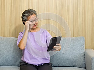Asian retired elderly with eyeglasses, sitting on the sofa watching at tablet computer. With presbyopia syndrome she cannot focus
