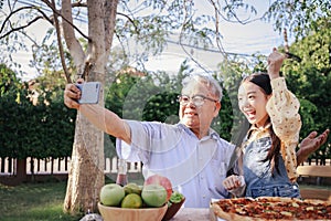 Asian retirement grandfather and pretty granddaughter enjoying to selfie while eating pizza together in home garden. Happy senior