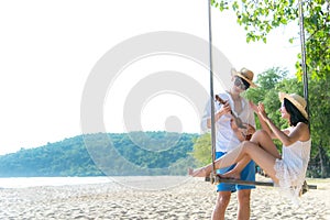 Asian romantic couple is sitting on sea beach on rope swing relax and happiness for holiday.  Honeymoon relax together on summer t