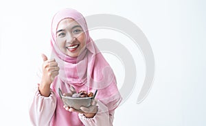 Asian 30s Muslim woman wearing traditional clothes with pink hijab headscarf, smiling holding a bowl of dry dates fruit in hands,