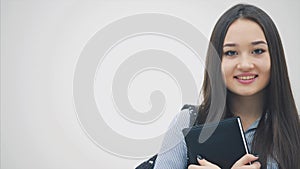 An asian schoolgirl appearing on a white background with a backpack slung over her shoulder, holding book near chest