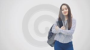 An asian schoolgirl appearing on a white background with a backpack slung over her shoulder, holding book near chest