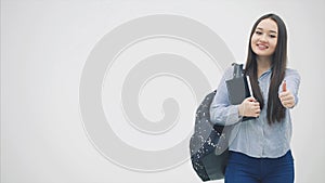 An asian schoolgirl standing with a backpack slung over her shoulder, holding book near chest, giving thumbs up.