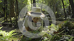 Asian schoolgirl wearing straw hat sitting and using magnifying glass to see the details of green plants and insects for education