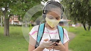Asian secondary schoolgirl in protective face mask wearing headphones and walking at public park in the residential area.
