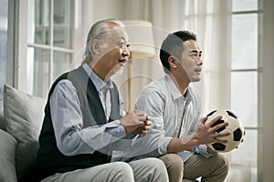 Asian seniior father and adult son watching football match on tv together