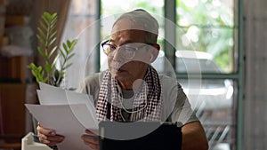 Asian senior businessman wears earphones in casual dress reading office document while using webinar on digital tablet at home.