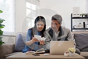 Asian senior couple checking and calculate financial bill together on sofa involved in financial paperwork, paying tax