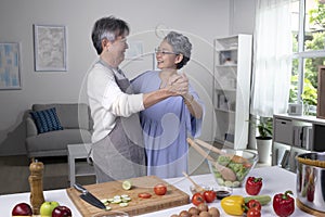 Asian senior couple is dancing and smiling while cooking together in kitchen