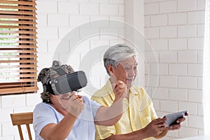 Asian senior couple play virtual realtiy glasses headset and tablet watching vr video and  have fun together in living room at