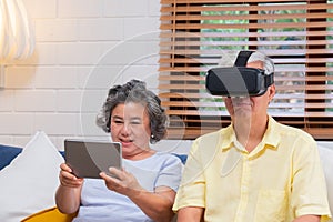 Asian senior couple play virtual realtiy glasses headset and tablet watching vr video and  have fun together on sofa  in living ro