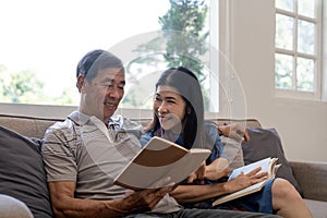 Asian senior couple reading book diary together, sitting on cozy sofa in home interior. Happy retirement activity
