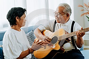 Asian senior couple sitting on the sofa and playing acoustic guitar together. Happy smiling elderly woman clapping hands