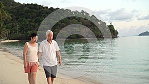 Asian senior elder couple walking holding hand together at tropical ocean