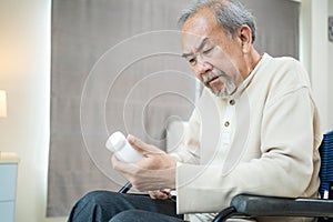 Asian Senior elder male patient sit on wheelchair at nursing home care. Mature older disabled man looking to medicine pill bottle