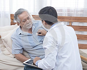 Asian senior elderly old man with mask on lying on sofa while young caucasian  doctor sit on knee check his heart beat and old