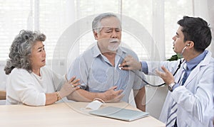 Asian senior elderly old man with mask on lying on sofa while young caucasian  doctor sit on knee check his heart beat and old
